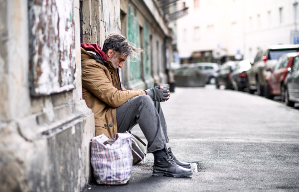 A side view of homeless beggar man sitting in front of wooden gate outdoors in city asking for money donation.