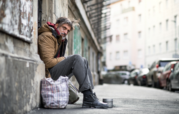 A homeless beggar man sitting in front of wooden gate outdoors in city, asking for money donation.