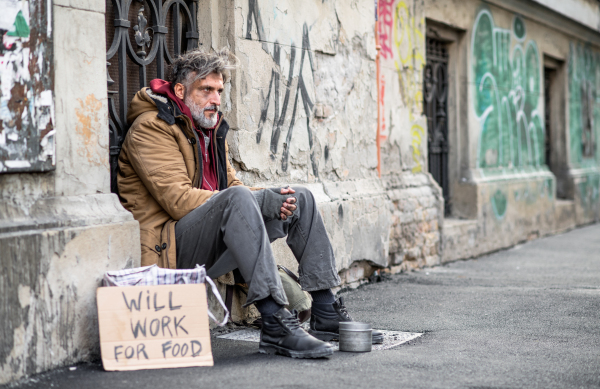 A side view of homeless beggar man sitting in front of gate outdoors in city, asking for money donation.