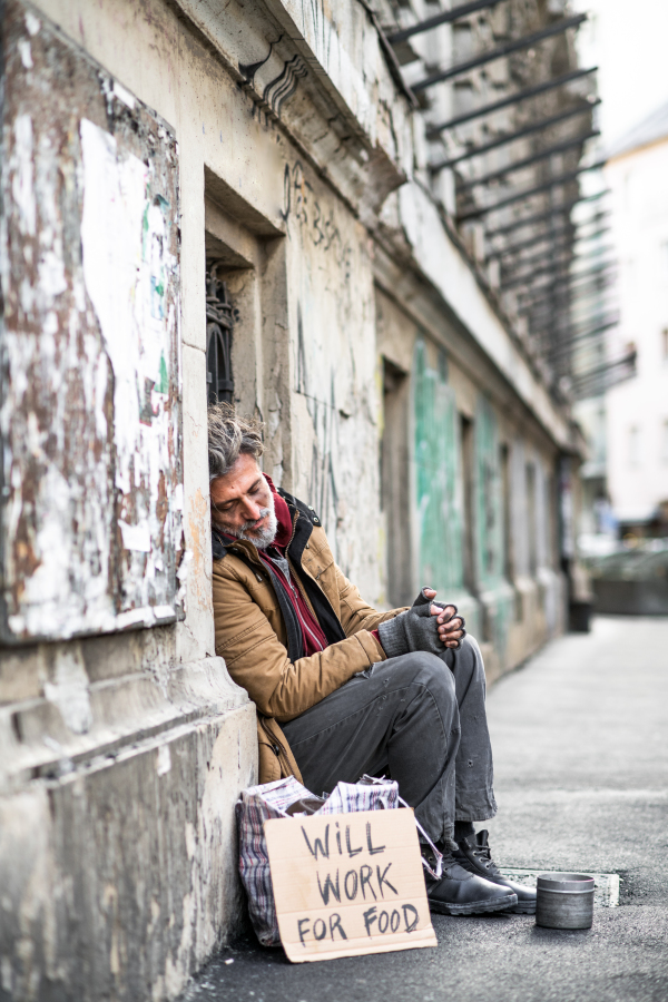 A side view of homeless beggar man sitting in front of wooden gate outdoors in city asking for money donation.