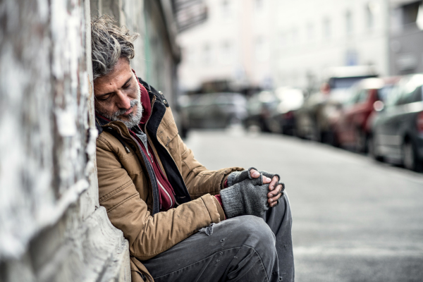 A homeless beggar man sitting in front of wooden gate outdoors in city asking for money donation, sleeping.