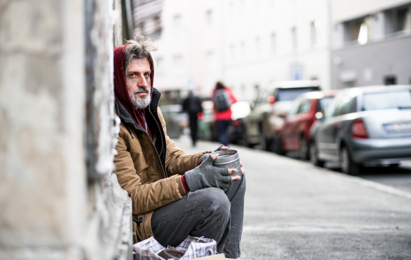 A side view of homeless beggar man sitting in front of wooden gate outdoors in city asking for money donation.