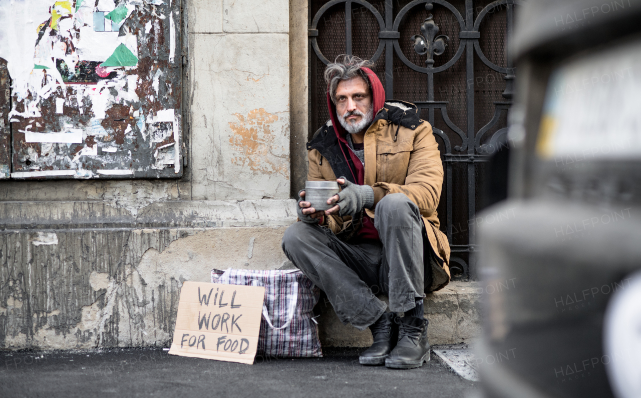 A homeless beggar man sitting in front of wooden gate outdoors in city asking for money donation.