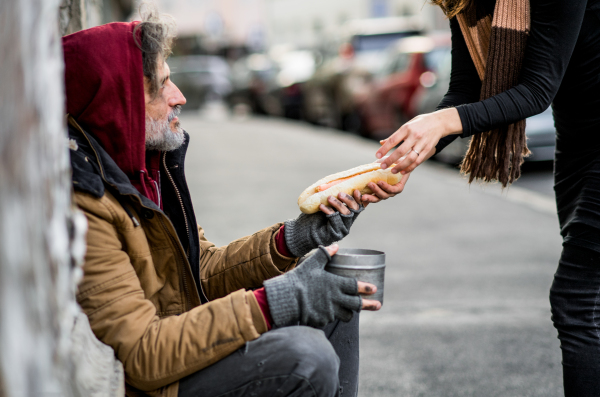 Unrecognizable woman giving food to homeless beggar man sitting outdoors in city.