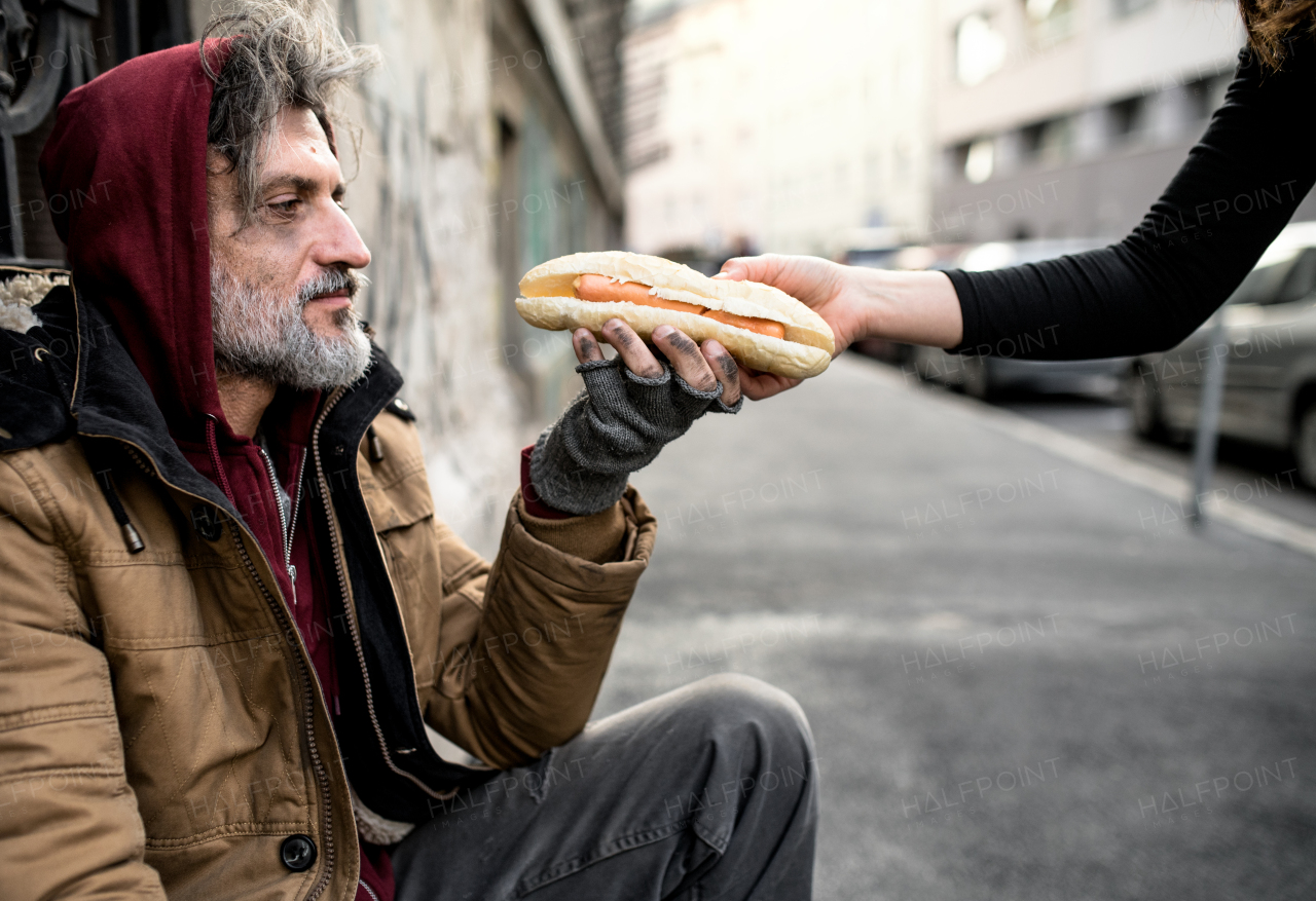 Unrecognizable woman giving food to homeless beggar man sitting outdoors in city.