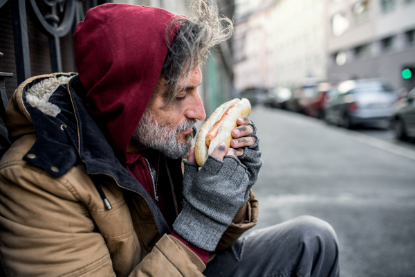 A homeless beggar man outdoors in city, holding and smelling hot-dog.