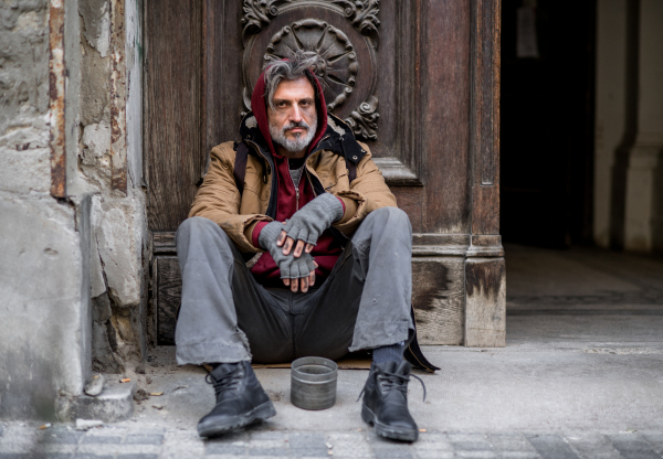 A homeless beggar man sitting in front of wooden gate outdoors in city asking for money donation.