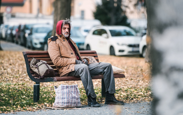 A homeless beggar man with a bag sitting on bench outdoors in city. Copy space.