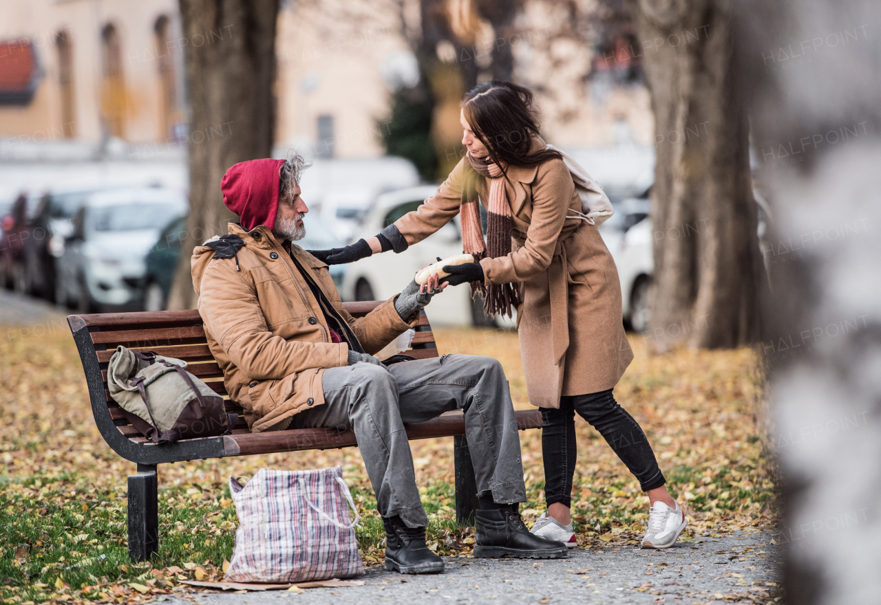 A young woman giving food to homeless beggar man sitting on a bench outdoors in city.