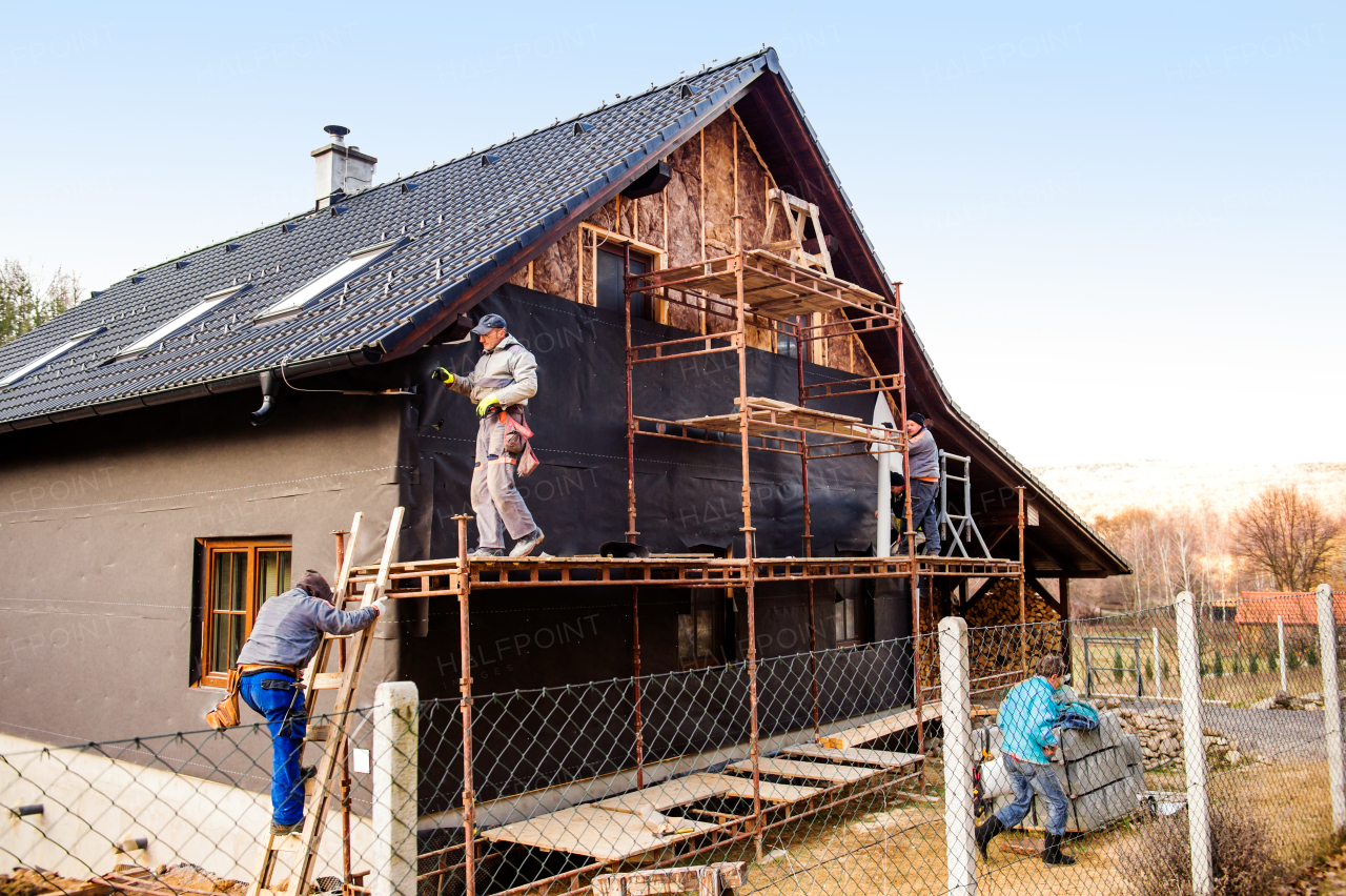 Construction workers standing on scaffold thermally insulating house facade with glass wool and black foil.