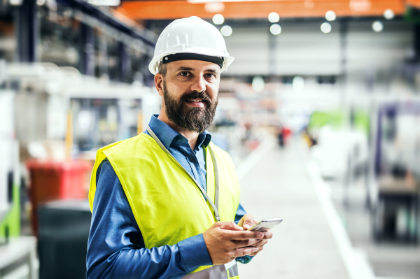 A mature industrial man engineer with name tag in a factory, using smartphone. Copy space.