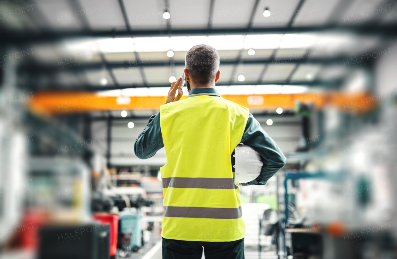 A rear view of a mature industrial man engineer with smartphone in a factory, making a phone call.