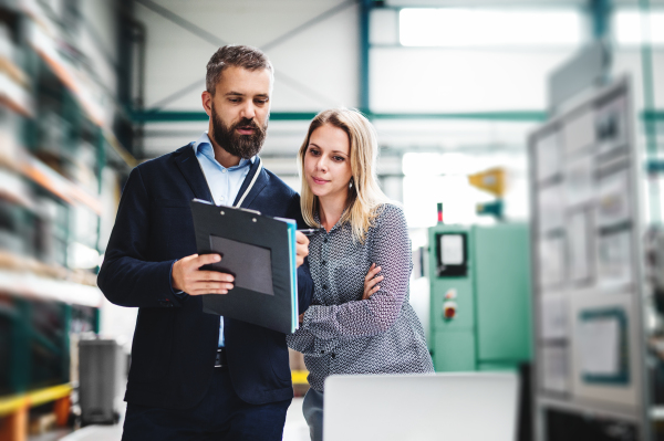 A portrait of a serious mature industrial man and woman engineer with laptop in a factory, working.