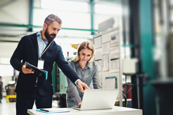 A portrait of a serious mature industrial man and woman engineer with laptop in a factory, working.