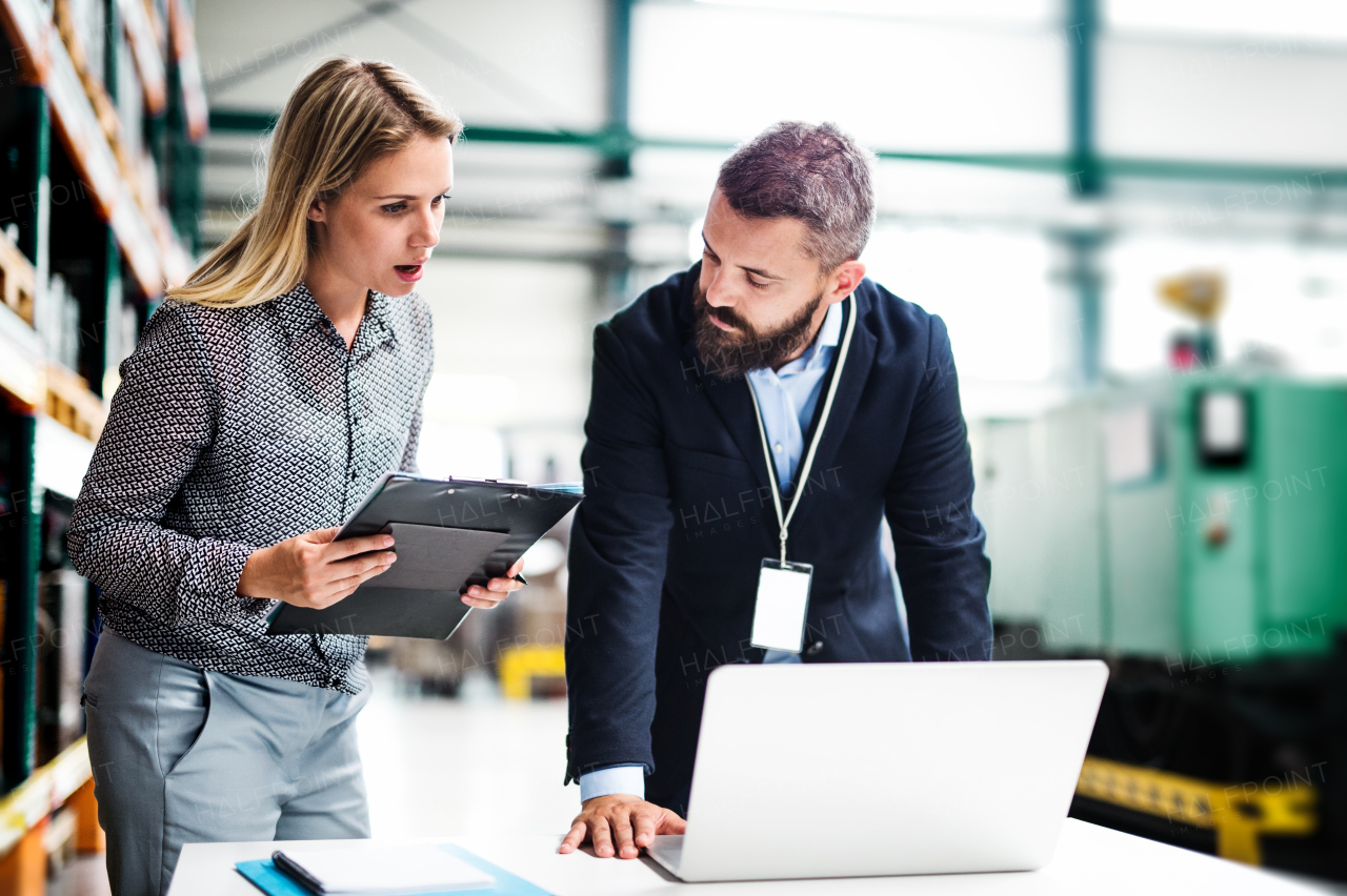 A portrait of a serious mature industrial man and woman engineer with laptop in a factory, working.