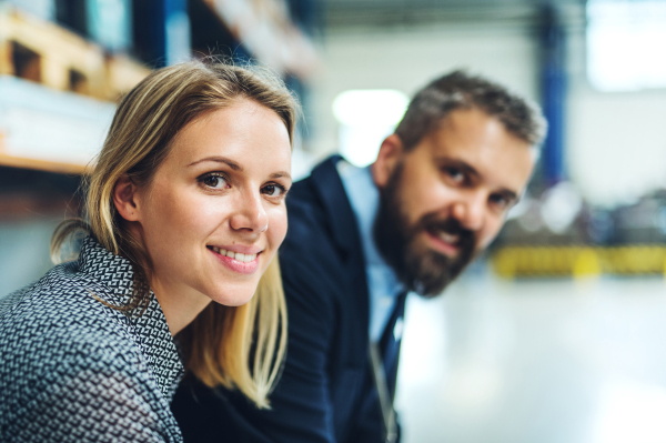 A portrait of a mature industrial man and young woman engineer in a factory, looking at camera.