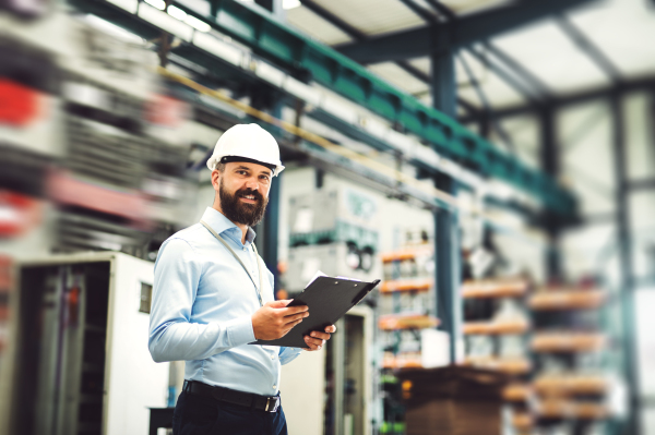 A portrait of a mature industrial man engineer with clipboard standing in a factory. Copy space.