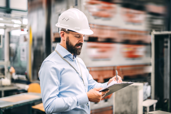 A portrait of a mature industrial man engineer with clipboard in a factory, writing. Copy space.