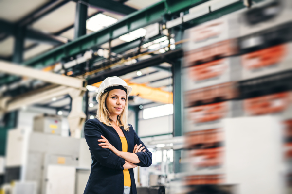 A portrait of a young industrial woman engineer standing in a factory, arms crossed.