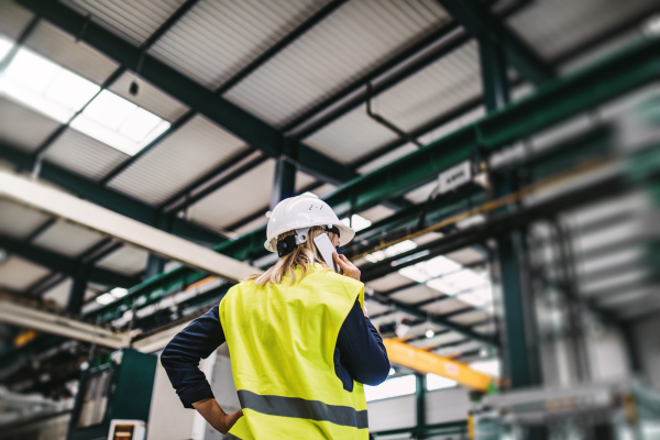 A rear view of a young industrial woman engineer with smartphone in a factory, making a phone call.