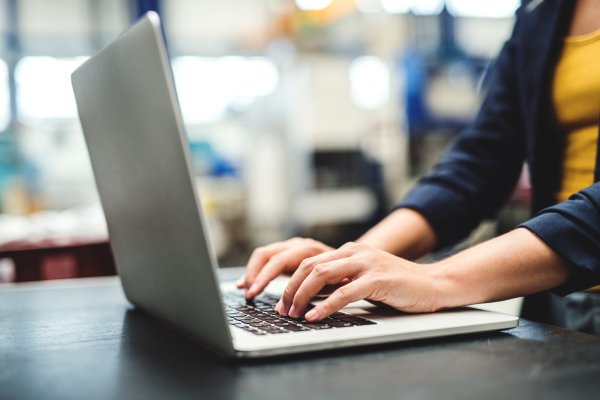 An unrecognizable industrial woman engineer in a factory with laptop sitting at the desk, typing.