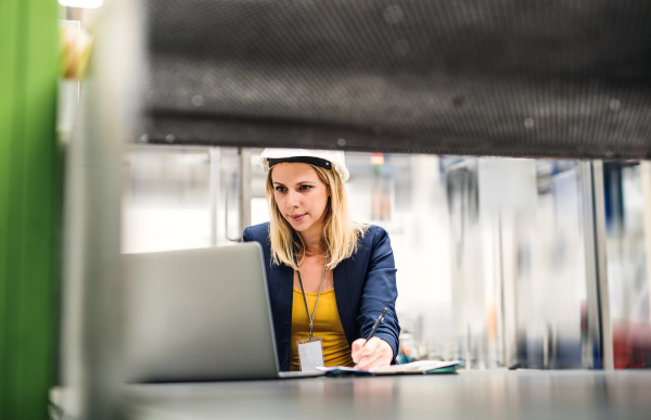 A portrait of a young industrial woman engineer in a factory, using laptop. Copy space.