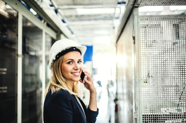 A portrait of a happy industrial woman engineer on the phone, standing in a factory.