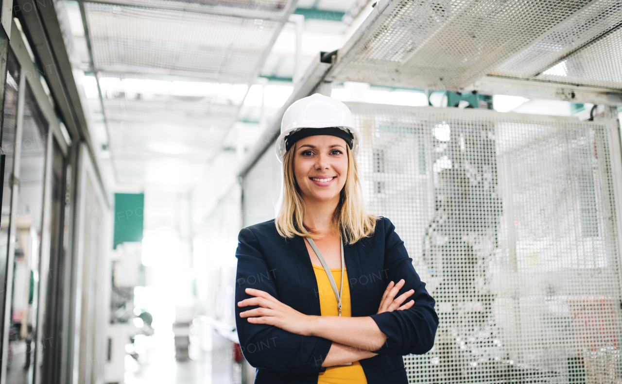 A portrait of a young industrial woman engineer standing in a factory, arms crossed.