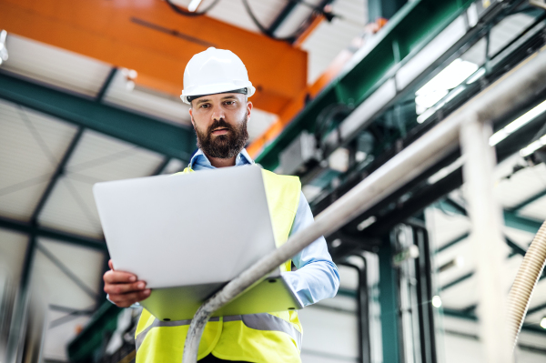 A portrait of a mature industrial man engineer with laptop in a factory, working.