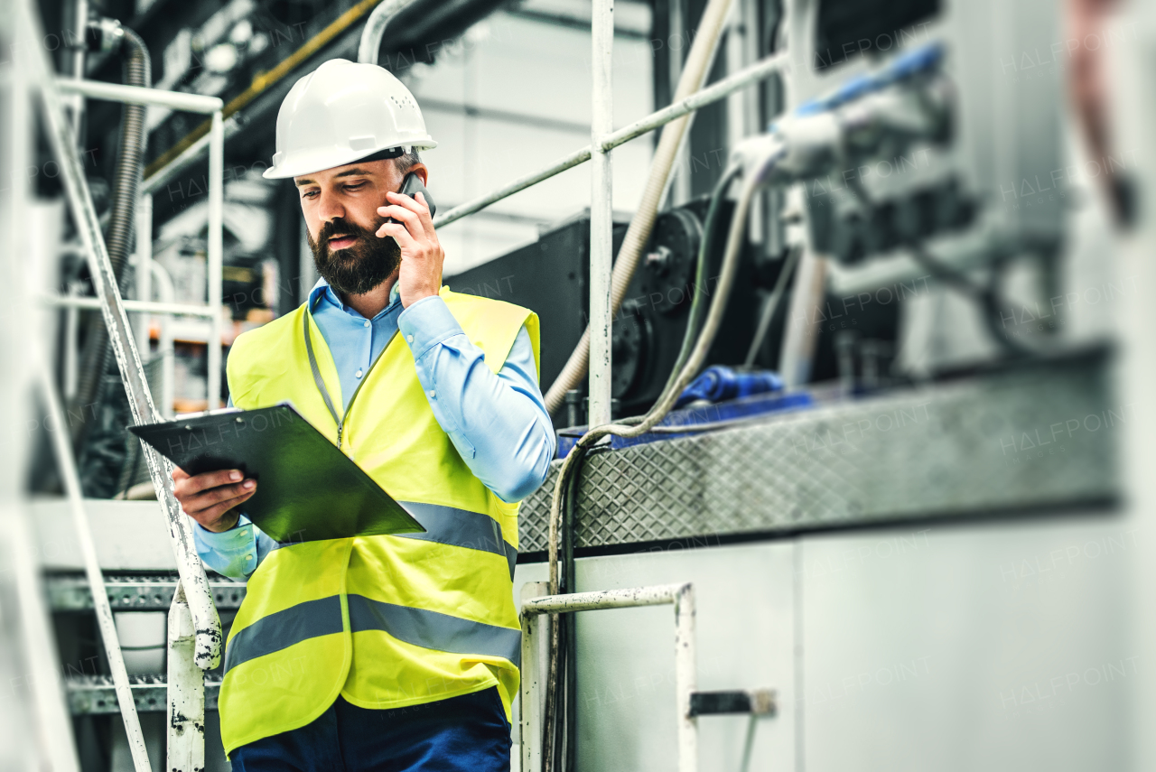 A portrait of a mature industrial man engineer with clipboard and smartphone in a factory, making a phone call. Copy space.