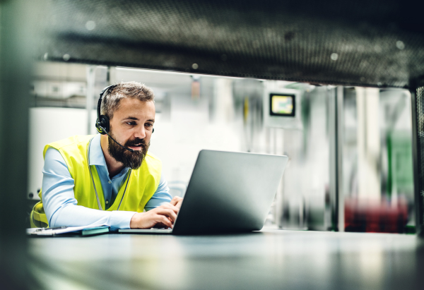 A portrait of a mature industrial man engineer with headset and laptop in a factory, working.