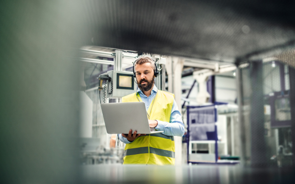 A portrait of a mature industrial man engineer with headset and laptop in a factory, working.