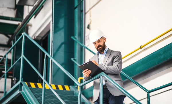 A portrait of a mature industrial man engineer with clipboard in a factory, writing. Copy space.