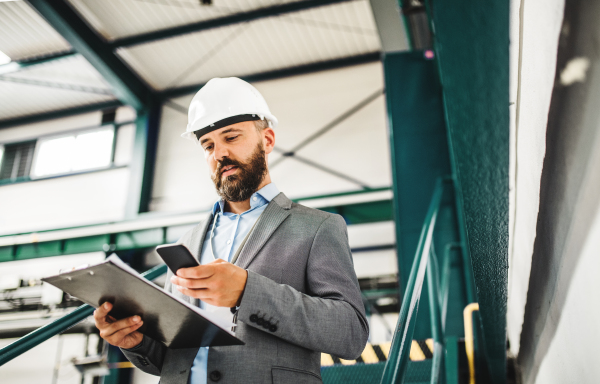 A portrait of a mature industrial man engineer with clipboard and smartphone in a factory, texting.