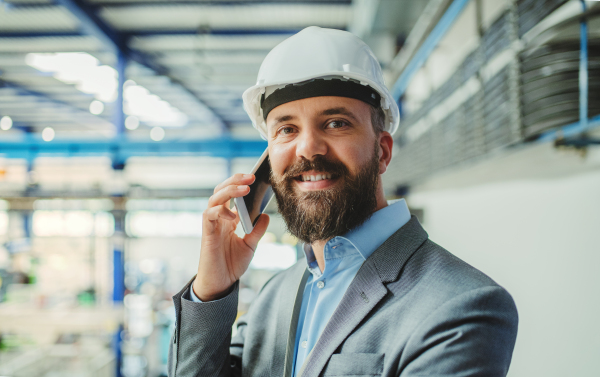 A portrait of a mature industrial man engineer with smartphone in a factory, making a phone call.