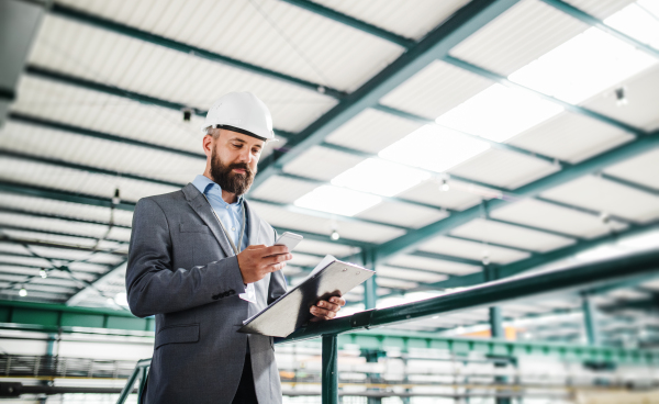 A portrait of a mature industrial man engineer with clipboard and smartphone in a factory, texting. Copy space.