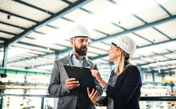 A portrait of a serious mature industrial man and woman engineer with clipboard in a factory, talking.