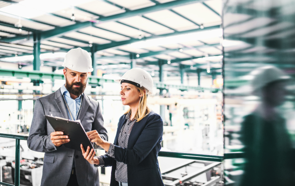 A portrait of a serious mature industrial man and woman engineer with clipboard in a factory, talking.