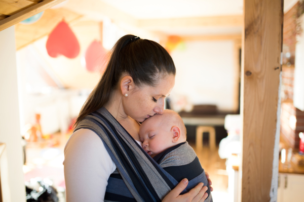 Beautiful young mother at home with her baby son sleeping in sling, kissing him on head