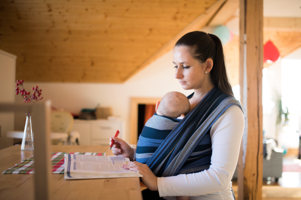 Beautiful young mother at home with her baby son sleeping in sling, reading a book or studying something