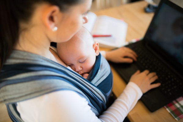 Unrecognizable young mother with her newborn baby son sleeping in sling at home, sitting at the table, writing on notebook keyboard