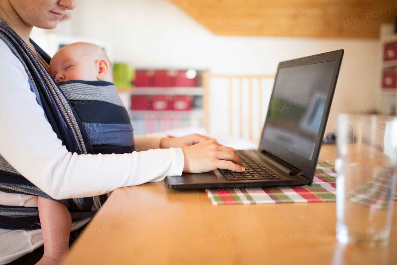 Unrecognizable young mother with her newborn baby son sleeping in sling at home, sitting at the table, writing on notebook keyboard