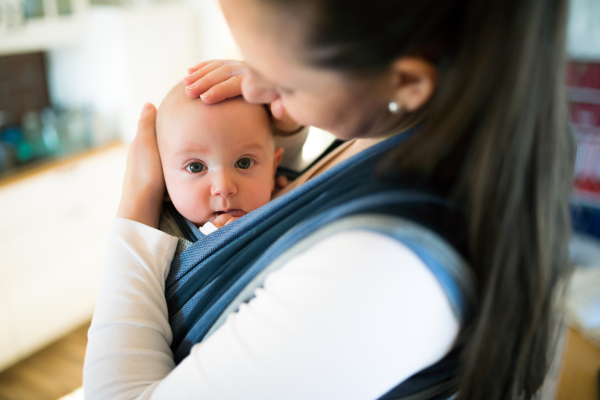 Beautiful young mother at home with her baby son in sling, stroking him gently