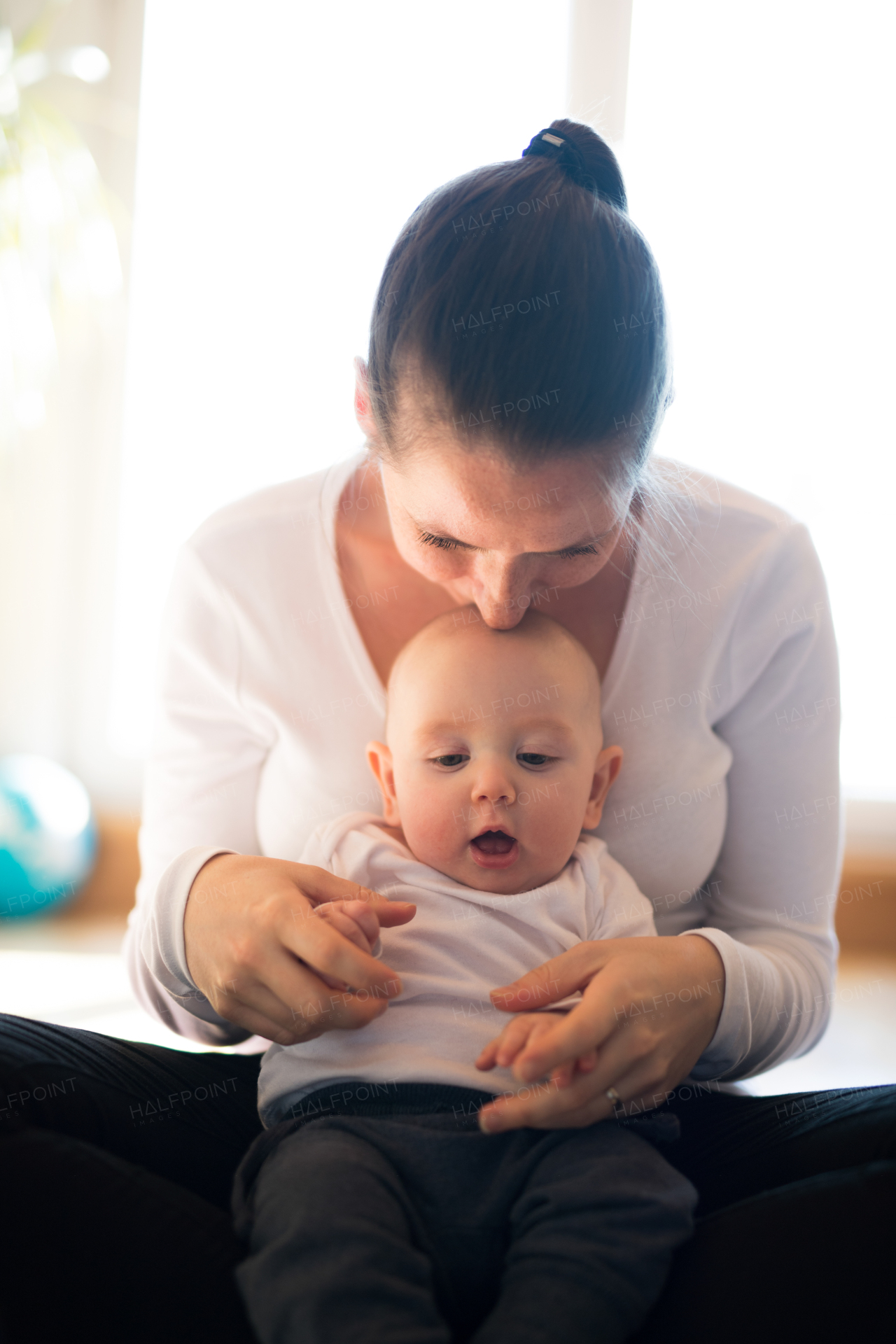 Beautiful mother in white t-shirt holding her cute baby son, sitting in her lap, kissing him.