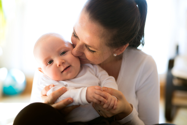 Beautiful mother in white t-shirt holding her cute baby son, sitting in her lap, kissing him.