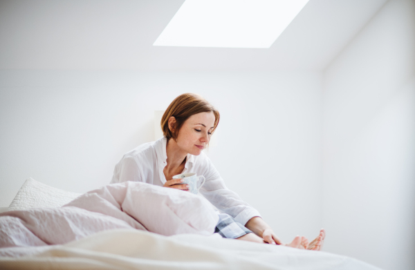 A young woman with night shirt sitting indoors on bed in the morning in a bedroom, holding a cup.