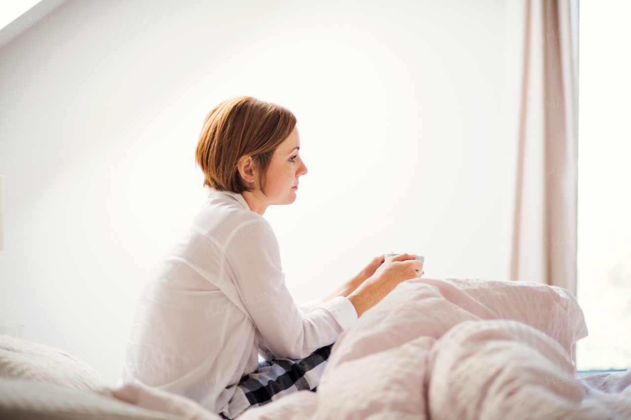 A young woman with night shirt sitting indoors on bed in the morning in a bedroom, holding cup.