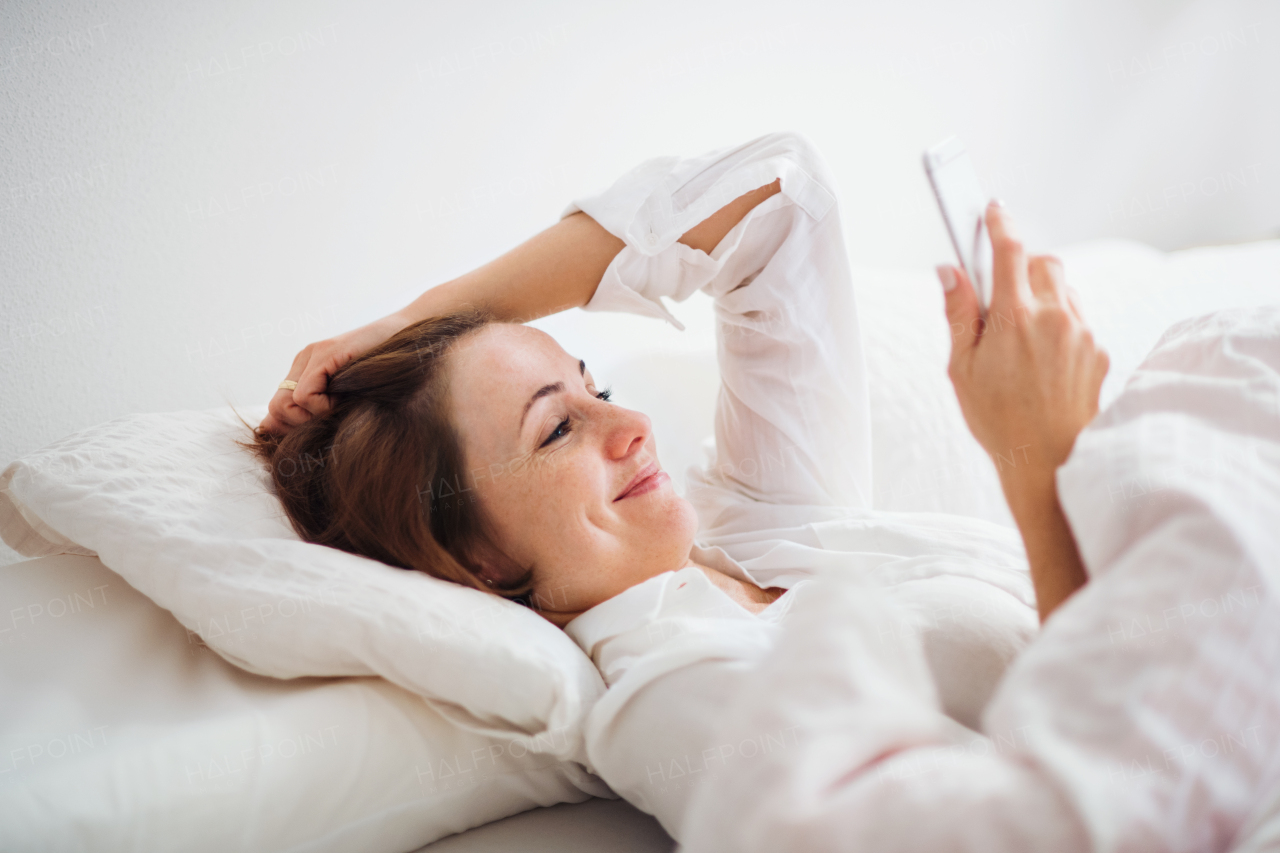 A happy young woman lying in bed indoors in the morning in a bedroom, using smartphone.