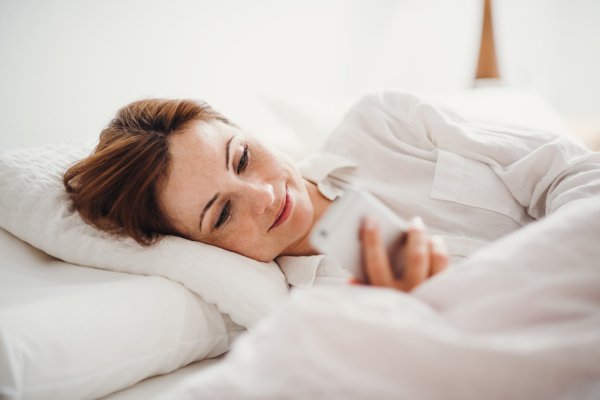 A happy young woman lying in bed indoors in the morning in a bedroom, using smartphone.