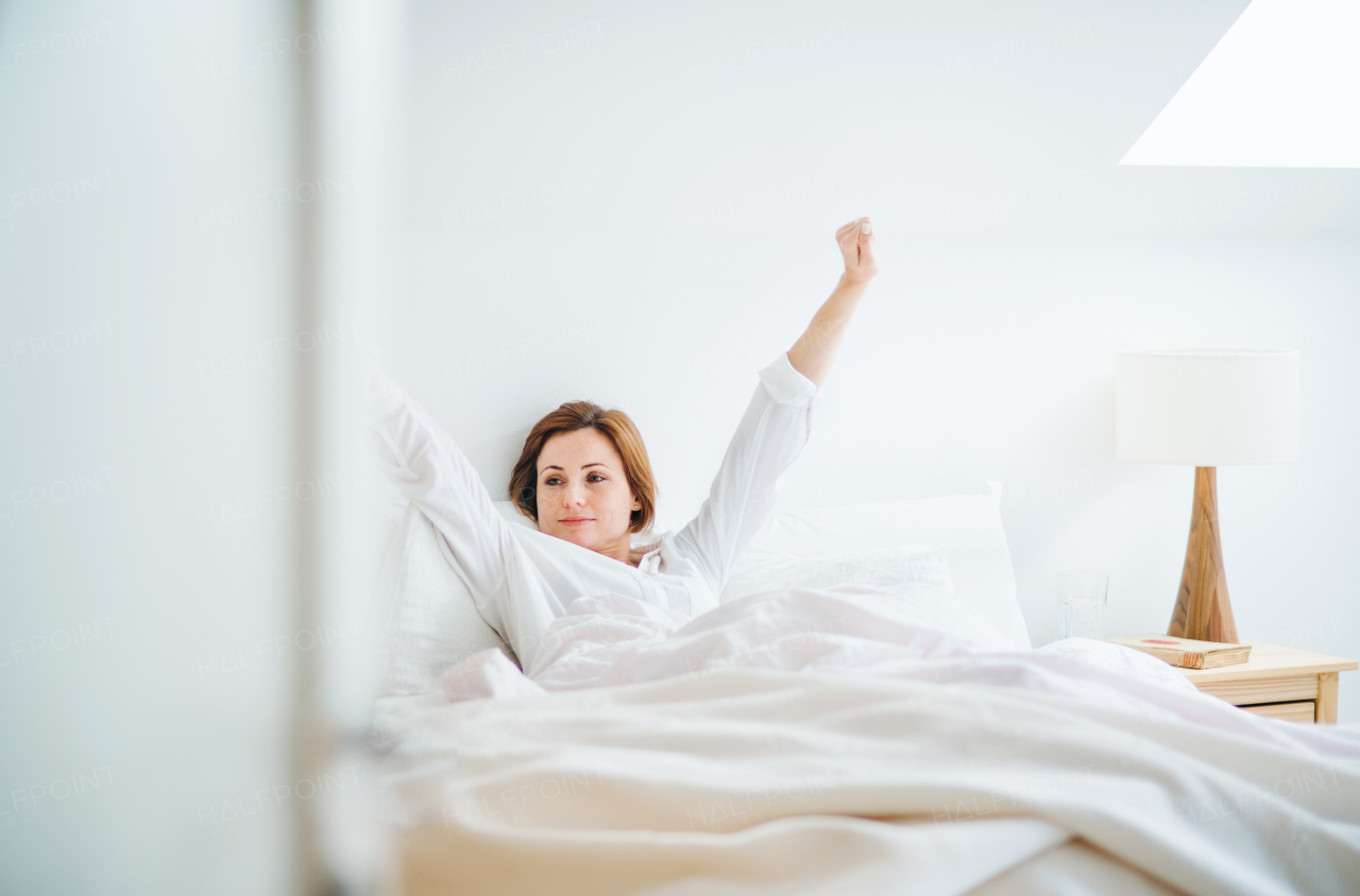 A happy young woman sitting in bed indoors in the morning in a bedroom, stretching.